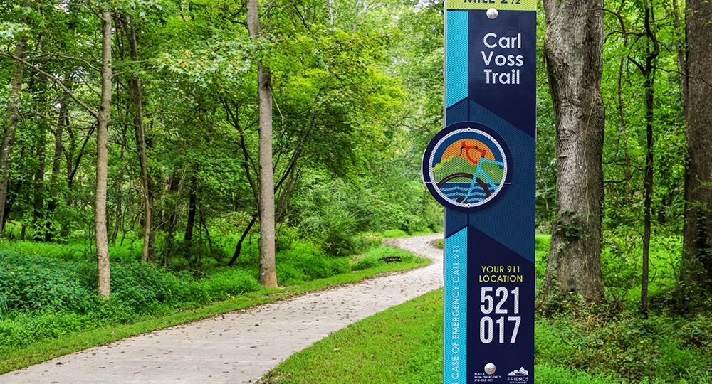 Carl Voss Trail E-911 sign amidst green trees beside a paved trail