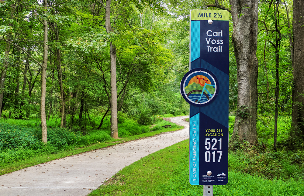 Carl Voss Trail E-911 sign amidst green trees beside a paved trail